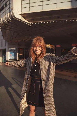 Stylish woman with red hair and a coat smiles in front of a theater entrance with poster displays, capturing a candid urban moment.