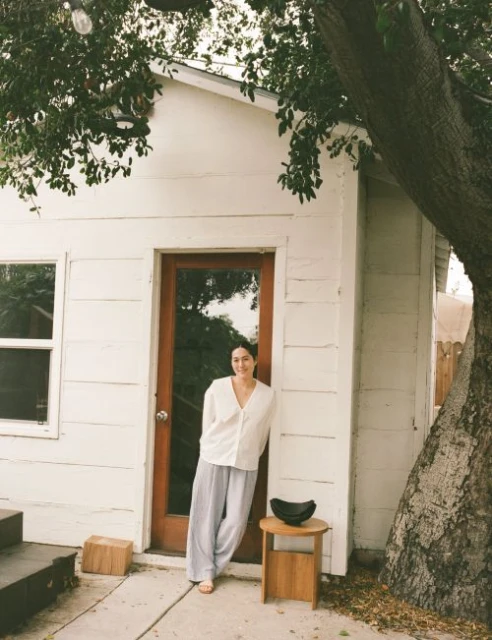 Woman in a white blouse and grey pants stands outside a white building with a brown door, next to a tree and small wooden table.