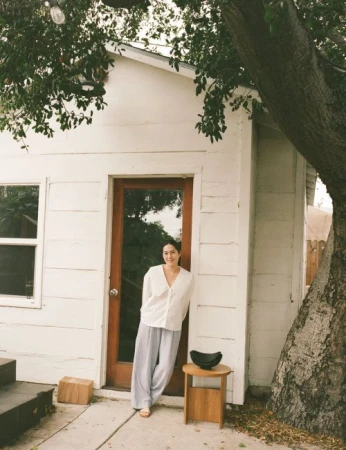 Woman in a white blouse and grey pants stands outside a white building with a brown door, next to a tree and small wooden table.