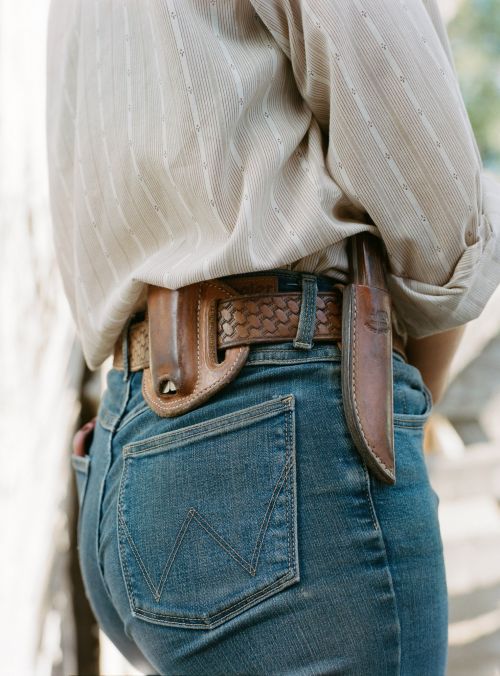 Woman with brown leather belt and knife sheath, wearing blue jeans and a striped shirt in an outdoor environment.