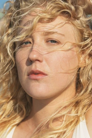 Close-up portrait of a woman with long blonde curly hair blowing across her face in natural sunlight.