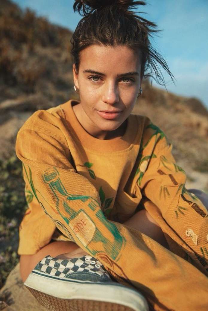 Portrait of a young woman with a messy bun, wearing a yellow graphic sweatshirt and checkered sneakers, posing outdoors in natural light.