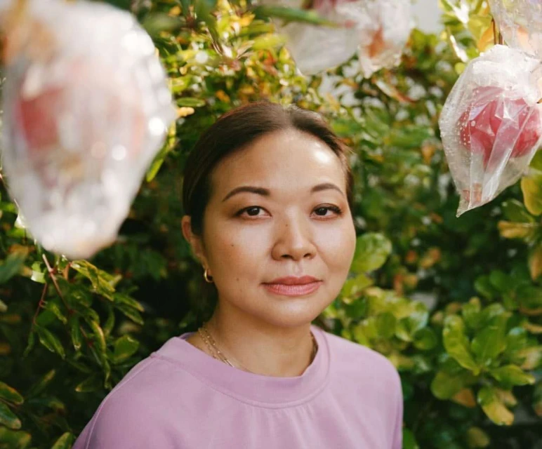 Close-up portrait of an Asian woman with dark hair, wearing a pink sweatshirt, standing outdoors among green foliage and fruit bags.