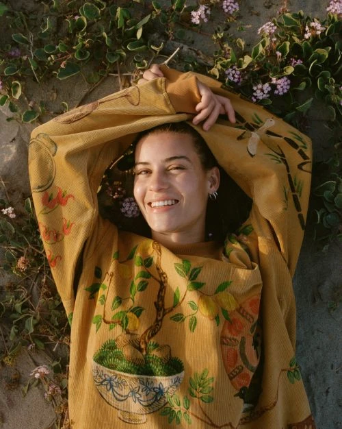 Smiling young woman lies in natural outdoor setting wearing a patterned sweatshirt, surrounded by plants and small purple flowers.