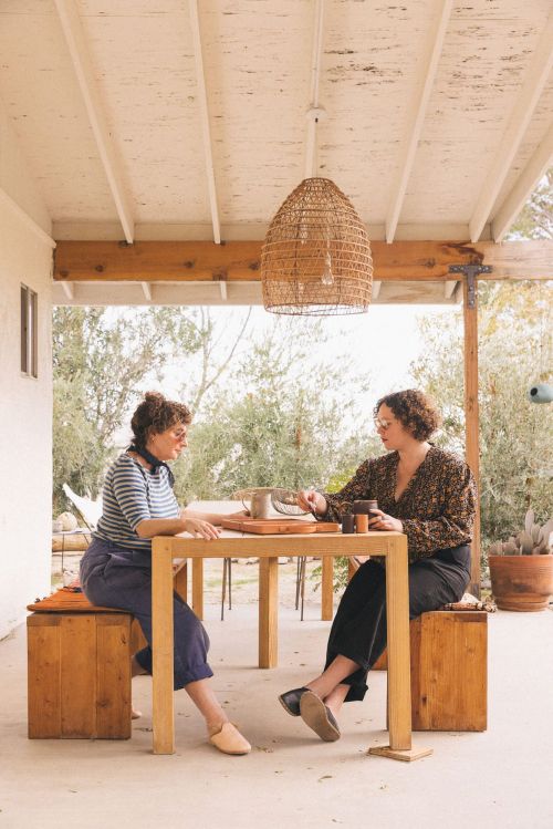 Two adult women sit at a wooden table on a porch under a woven light fixture, engaged in a relaxed conversation in natural daylight.