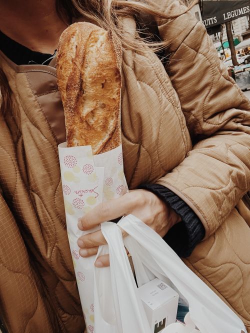 Person in a tan quilted jacket holds a wrapped baguette and shopping bag in a street scene with fruit and vegetable stand.