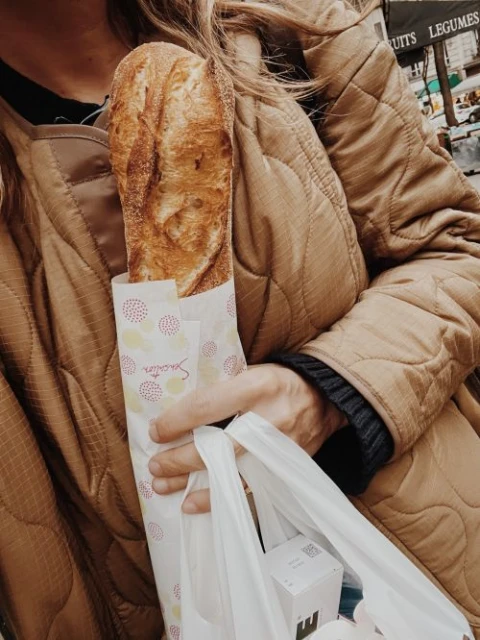 Person in a tan quilted jacket holds a wrapped baguette and shopping bag in a street scene with fruit and vegetable stand.