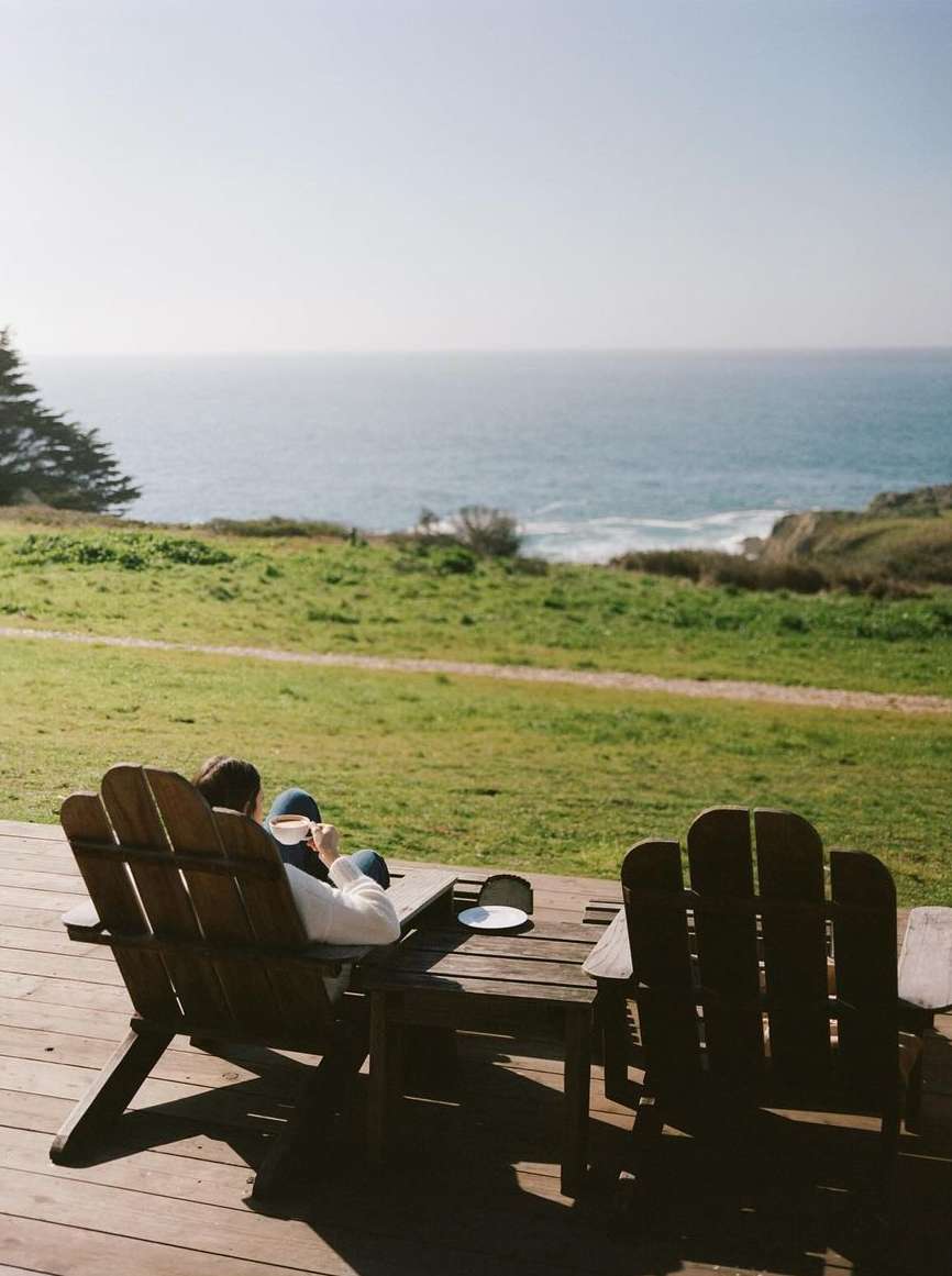 Person relaxing in an Adirondack chair on a porch overlooking the ocean and green landscape on a sunny day with clear skies and open space.