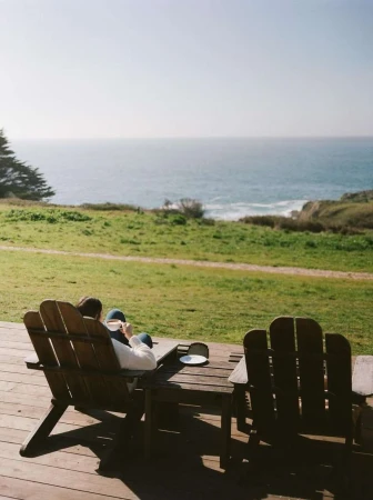 Person relaxing in an Adirondack chair on a porch overlooking the ocean and green landscape on a sunny day with clear skies and open space.