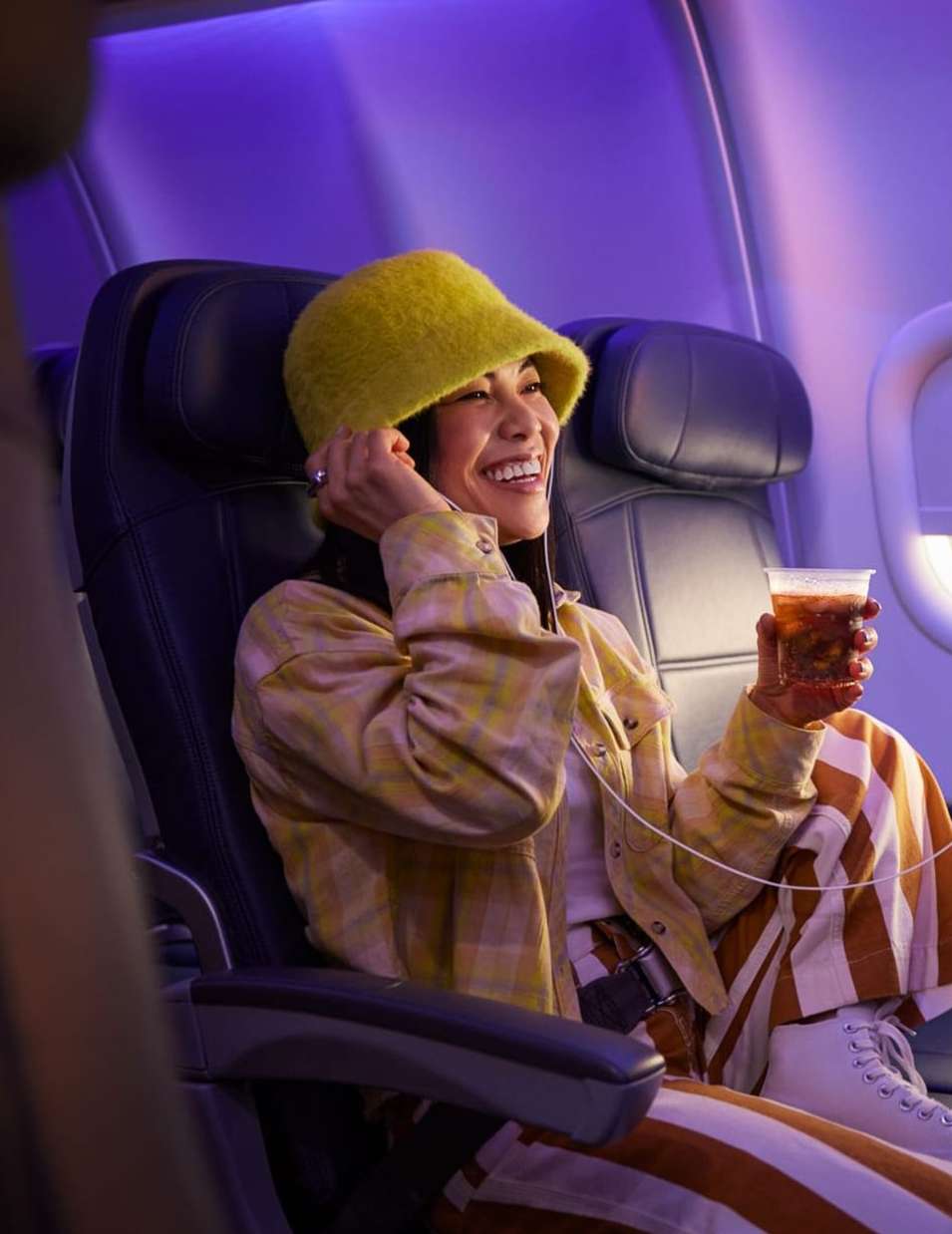 A smiling woman with a yellow bucket hat sits in an airplane seat holding a drink. She is wearing a yellow plaid jacket and striped pants.