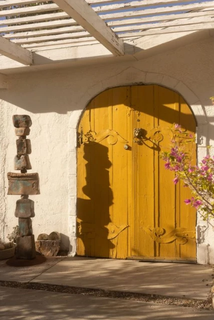 Exterior shot of a yellow wooden door with decorative hardware, flanked by a stone sculpture and a bougainvillea plant under a pergola.