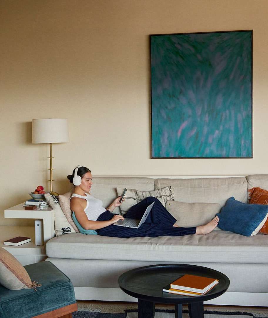 Woman wearing white headphones and a tank top seated on a beige couch with a laptop and phone by Anais and Dax