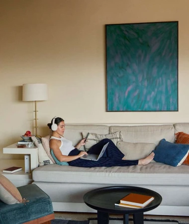 Woman wearing white headphones and a tank top seated on a beige couch with a laptop and phone by Anais and Dax