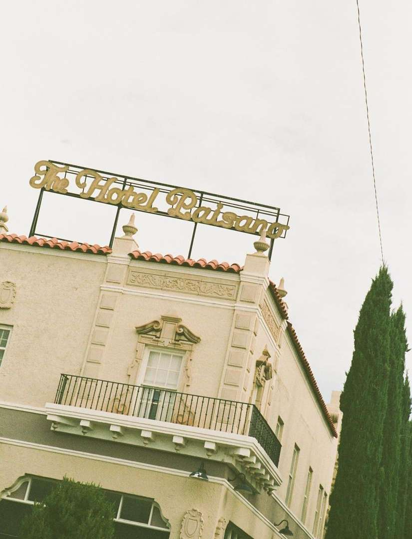 Exterior of The Hotel Paisano building with its distinctive gold sign on top, set against a cloudy sky and green trees.