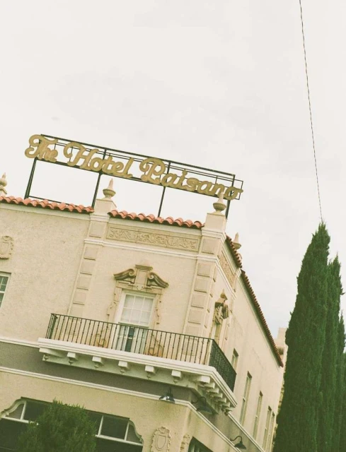 Exterior of The Hotel Paisano building with its distinctive gold sign on top, set against a cloudy sky and green trees.