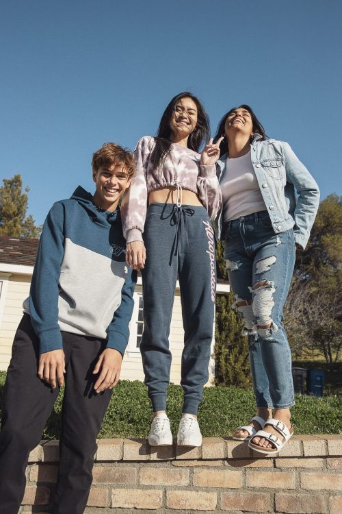 Three young adults, two women and one man, pose outdoors wearing casual clothing, standing on a brick wall on a sunny day.