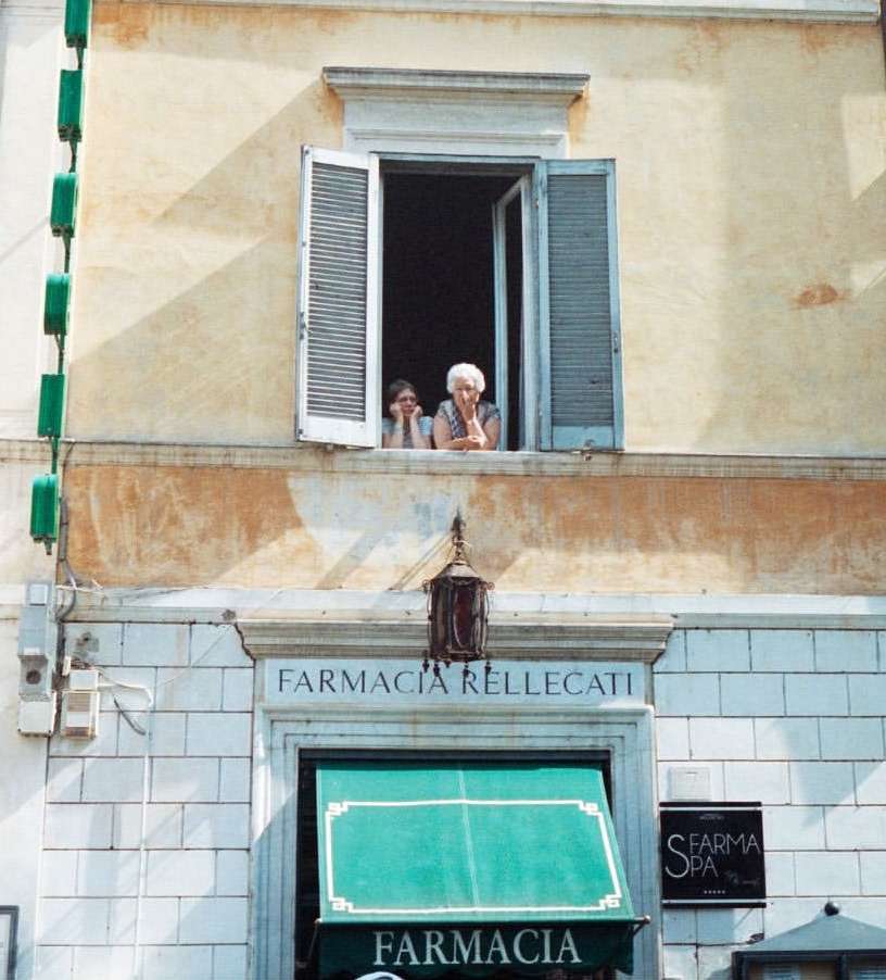 Two women are visible from a window above a pharmacy with cream and tan walls, complemented by green awnings and clear outdoor lighting.