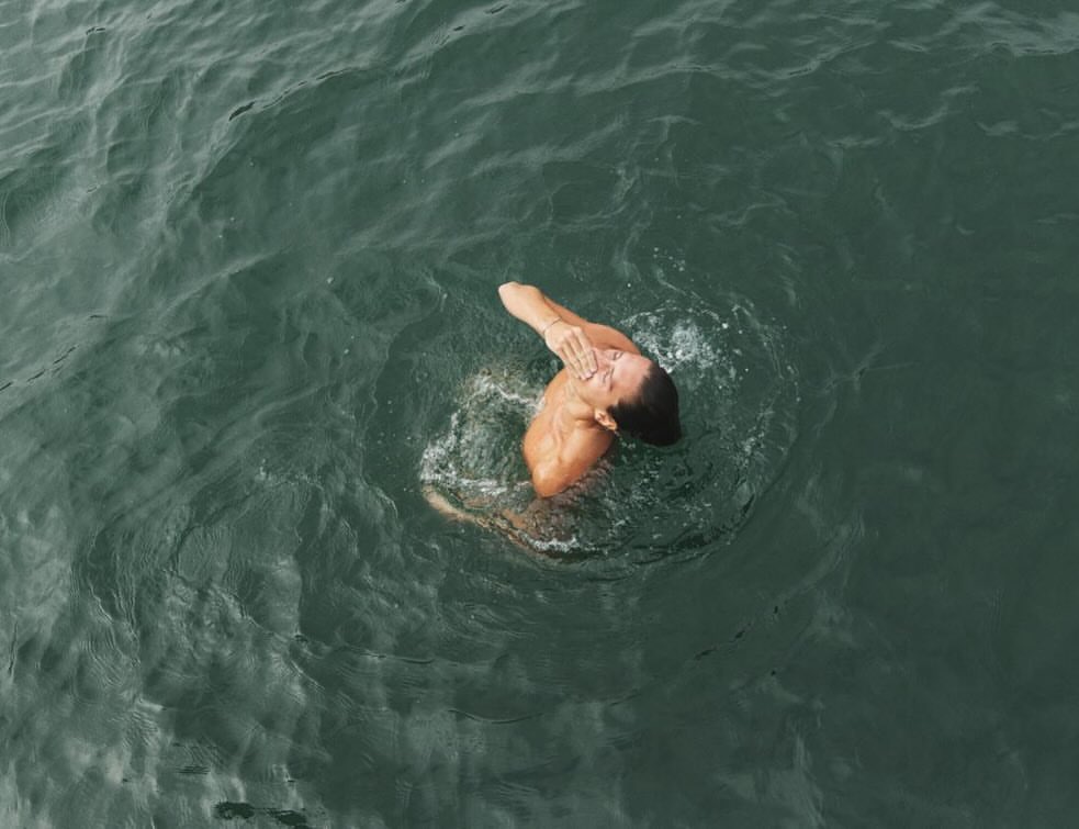 Overhead aerial shot of a person swimming in dark green ocean water, splashing and wiping their face with one hand.