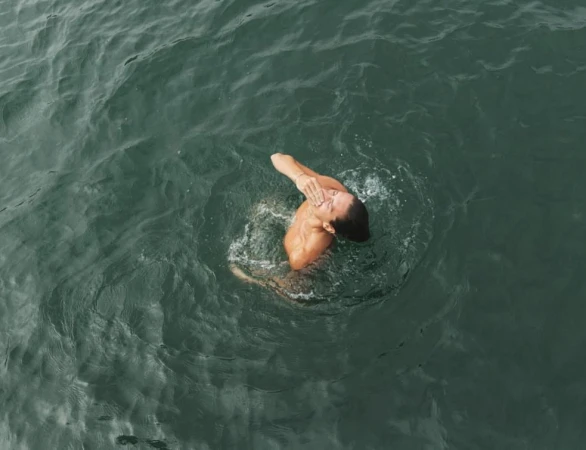 Overhead aerial shot of a person swimming in dark green ocean water, splashing and wiping their face with one hand.