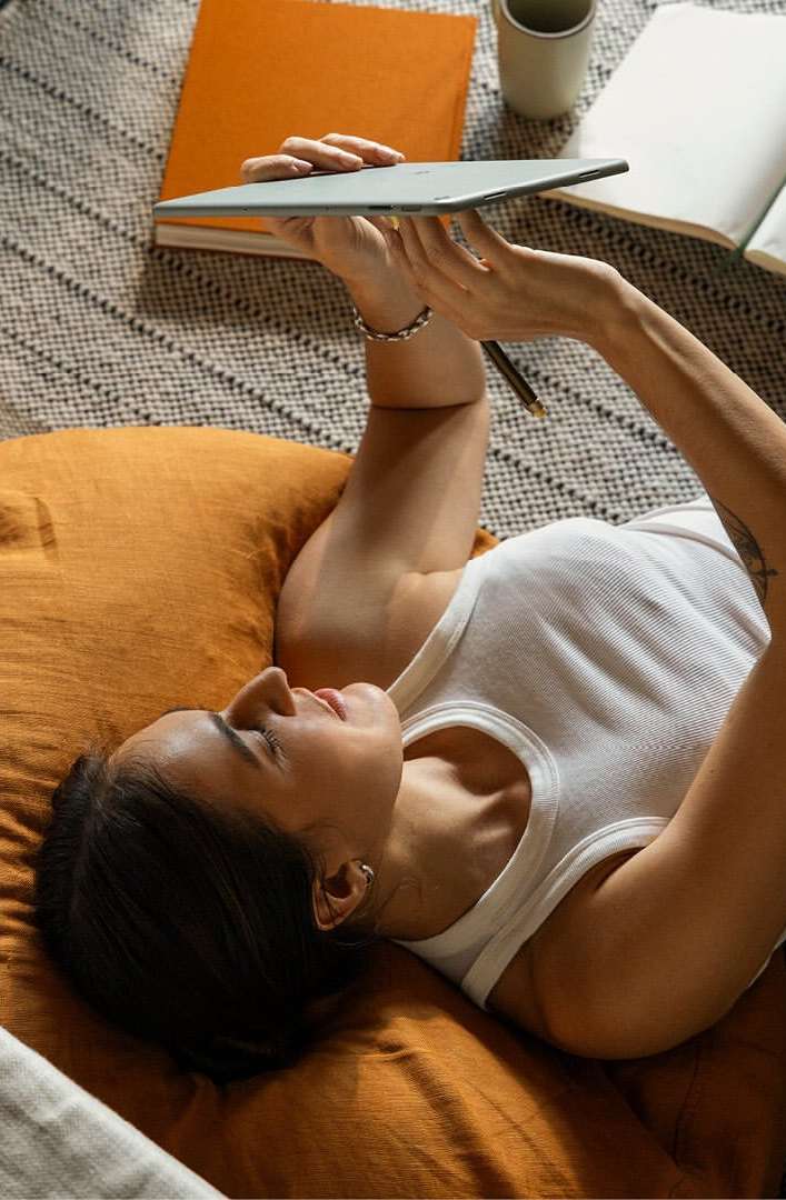 Woman lying on a cushion using a tablet device, with books and a mug in the background, creating a cozy and relaxed indoor scene.