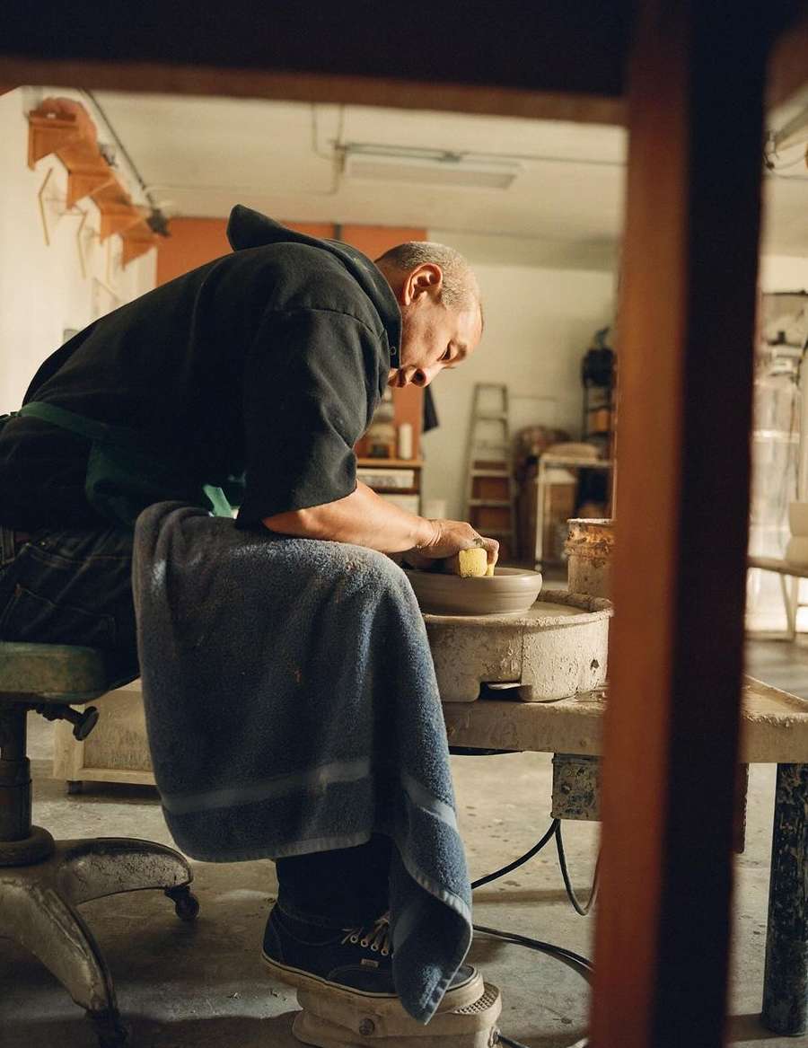 Adult male potter sitting at a pottery wheel in a studio, shaping clay.  The potter is wearing a dark hooded sweater and apron.