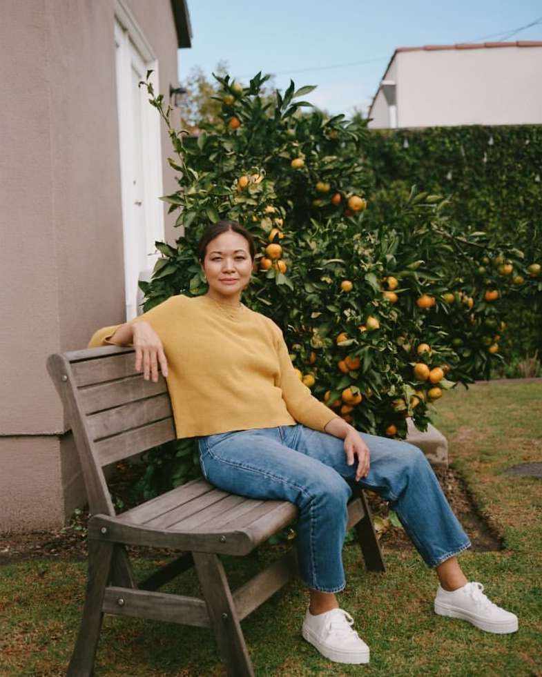Woman sitting on a wooden bench in a garden with an orange tree, wearing a yellow sweater, blue jeans, and white sneakers in daylight.