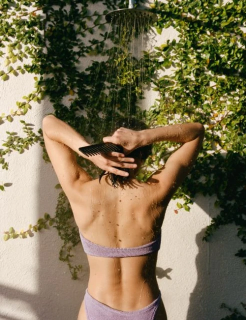Back view of a woman in a bikini showering outdoors while combing her hair, against a wall covered with green climbing vines.