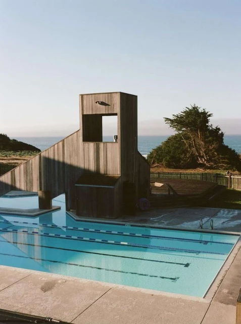 Outdoor swimming pool with blue water reflecting the sky, adjacent to a wooden structure and grassy area with ocean views.