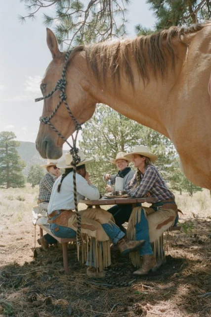 A group of people in cowboy attire sit at a picnic table in a field with a light-brown horse standing behind them in daylight.
