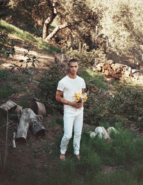 A bare-footed man in a white outfit stands in a sunlit, grassy clearing, holding a small bouquet of yellow flowers, surrounded by trees.