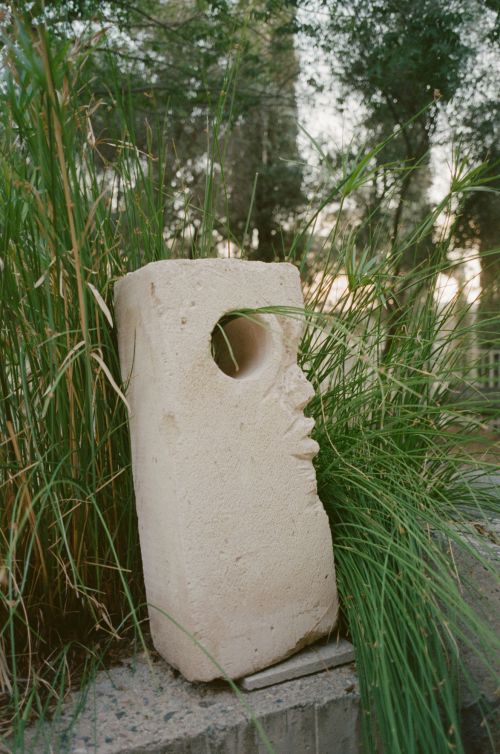 Stone sculpture stands outdoors among green grasses and trees, featuring a hollowed-out round hole, creating an abstract art piece.