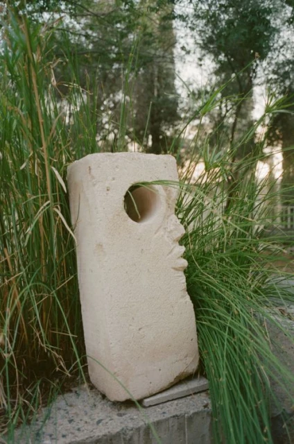 Stone sculpture stands outdoors among green grasses and trees, featuring a hollowed-out round hole, creating an abstract art piece.