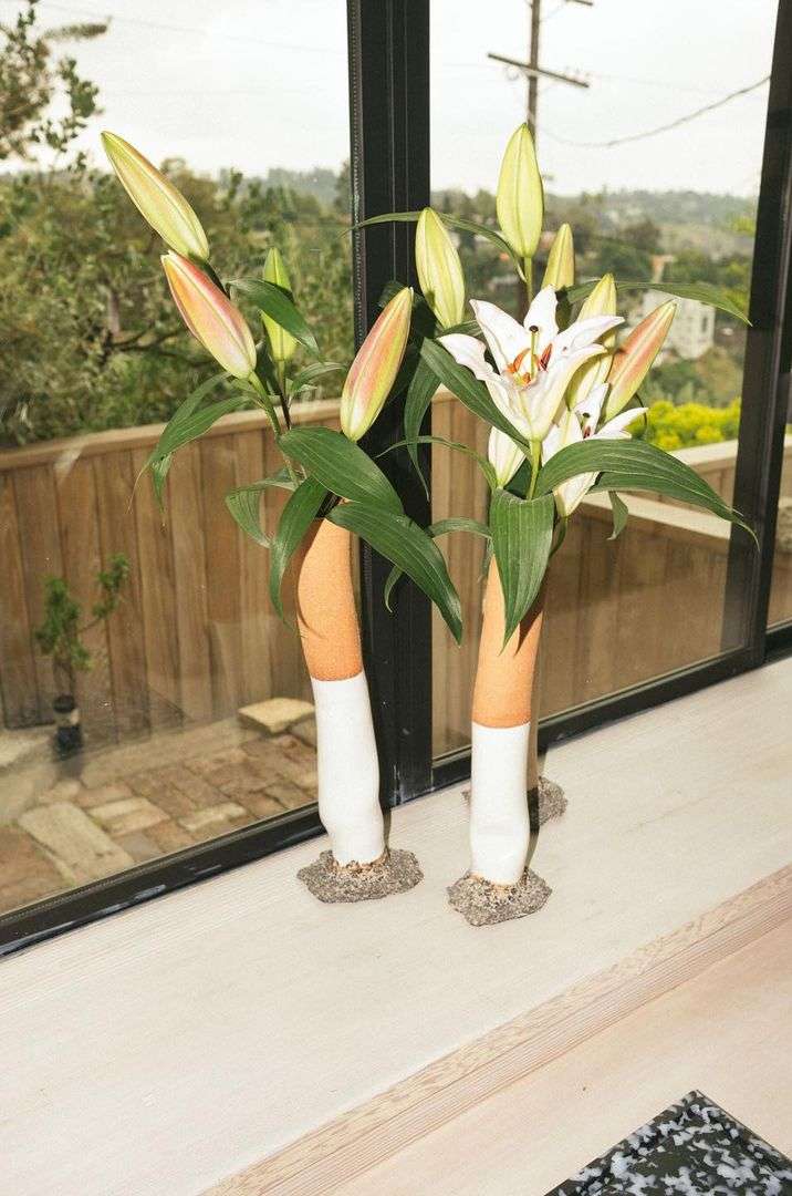 Still life shot of two ceramic vases holding lilies on a white windowsill, with an out-of-focus outdoor scene visible through a window.