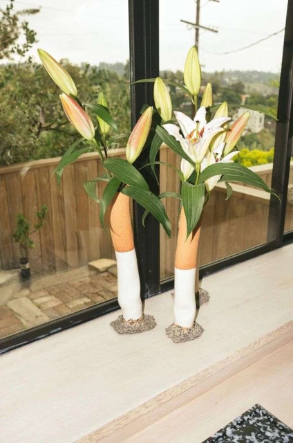 Still life shot of two ceramic vases holding lilies on a white windowsill, with an out-of-focus outdoor scene visible through a window.