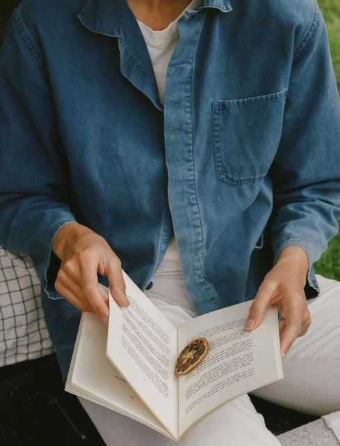 Person in blue denim jacket reading a book with a dried orange slice on the page, sitting outdoors on a checkered surface.