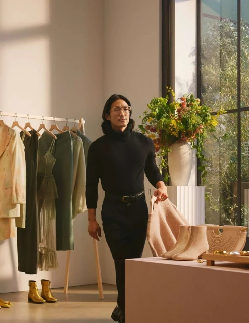 A stylish man in a black turtleneck holds a sweater next to a table with boots and a purse, clothes hang from a rack in the background.