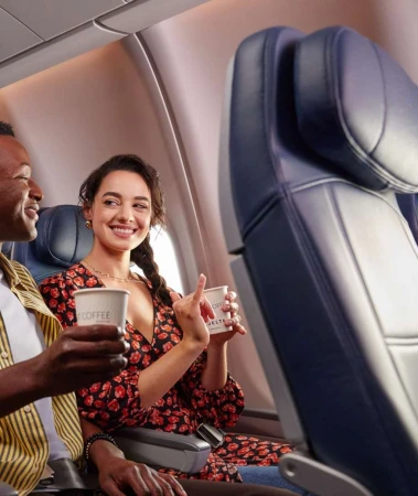 Two smiling passengers on an airplane, a Black man and a White woman, holding paper coffee cups near a window seat.