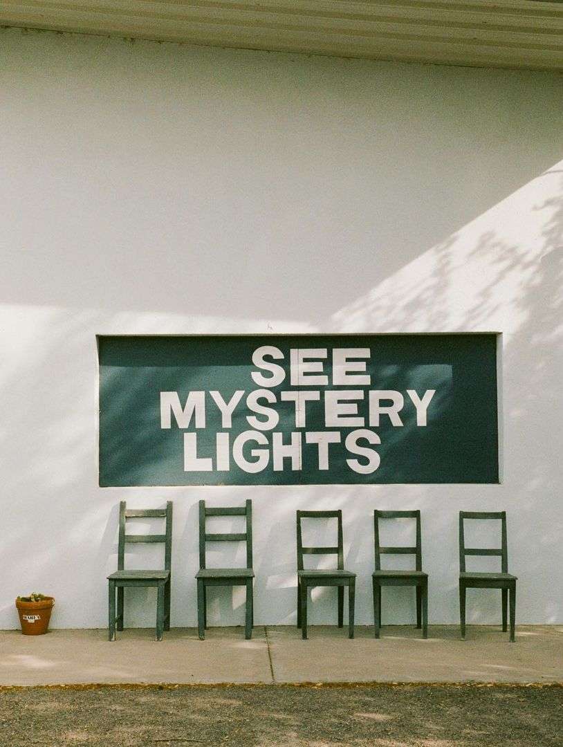 Row of chairs in front of a white wall with a sign that reads "See Mystery Lights", casting shadows of tree branches.