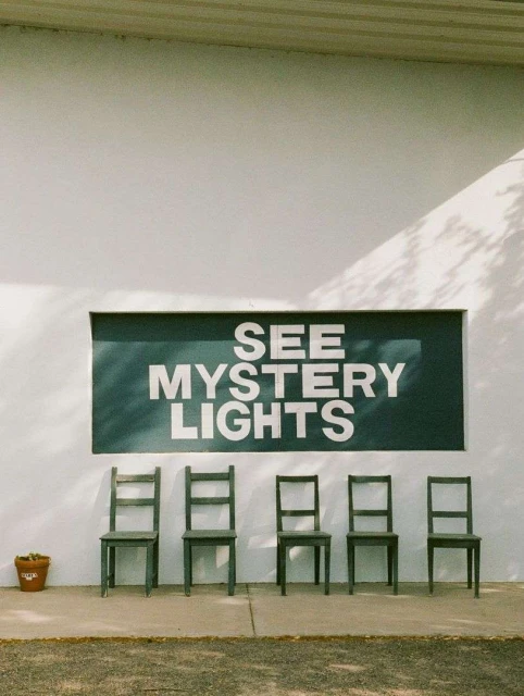 Row of chairs in front of a white wall with a sign that reads "See Mystery Lights", casting shadows of tree branches.