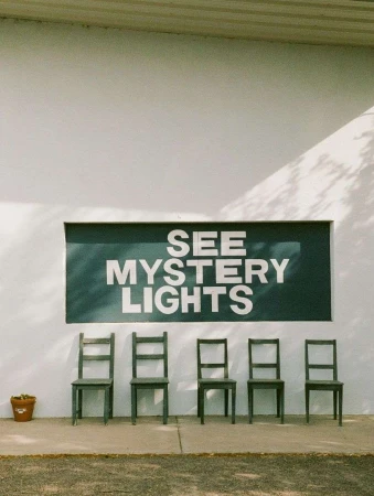 Row of chairs in front of a white wall with a sign that reads "See Mystery Lights", casting shadows of tree branches.