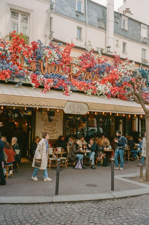 Outdoor cafe in Paris with people sitting at tables, vibrant red, white, and blue floral decorations, and cobblestone street.