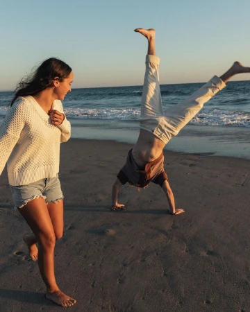 Woman in white sweater smiling as another person does a handstand on a sandy beach at sunset.