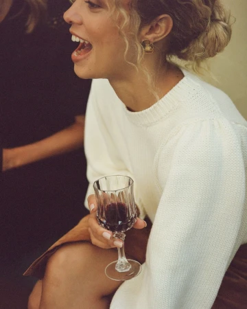 Smiling woman in a white sweater holding a crystal glass of red wine, photographed by Anais and Dax.
