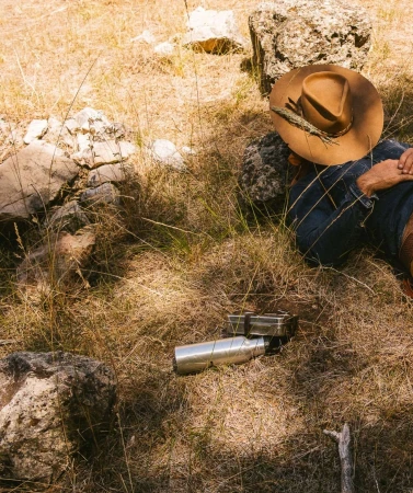 Man wearing a brown cowboy hat and denim shirt resting among rocks and dry grass with a metal water bottle nearby.