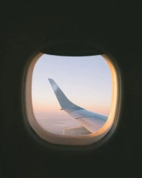 Airplane wing viewed through an oval cabin window at sunset with soft pastel sky colours.