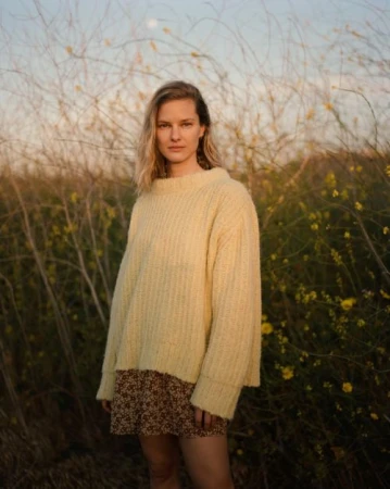 A woman wearing a pale yellow knit sweater and a patterned skirt standing outdoors among tall dry weeds and small yellow wildflowers at dusk.