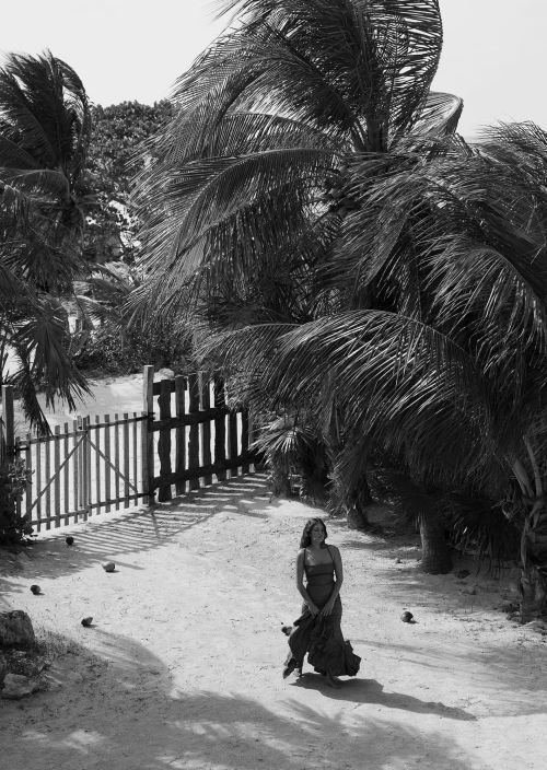 Black and white portrait of a woman standing on a sandy path near a gate and palm trees, captured in an outdoor setting.