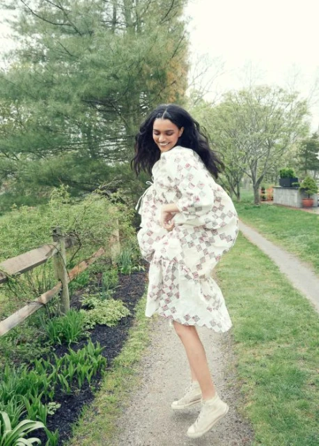 A cheerful woman with long dark hair jumps on a gravel path while wearing a white patterned dress in an outdoor park setting.