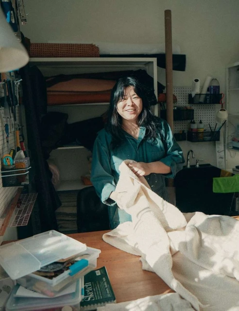 Smiling Asian woman in a green shirt holds beige fabric in an indoor workshop with craft supplies and shelves filled with material.