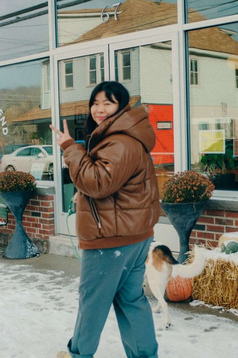 Asian woman in brown puffer jacket flashes peace sign in snowy urban setting with dog, plants, buildings reflected in window.